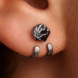 May include: Close-up of a dark gray earring set. The set includes a textured, round stud and a curved, open-hoop earring. The jewelry is worn on an ear, with the focus on the earring design and placement.