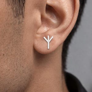 以下が含まれることがあります： アルギズのルーン文字の形をした小さなシルバーカラーのスタッドピアス。垂直の線と上向きに伸びる2本の枝で構成されています。滑らかで光沢のある金属仕上げで、耳たぶにしっかりと装着されています。