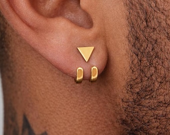 May include: Close-up of gold earrings. One earring features a triangle shape, while the other has two small, connected, oval shapes. The earrings are displayed on an ear, showcasing a minimalist jewelry design.
