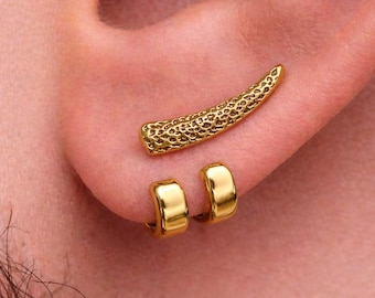 May include: Gold-colored ear jewelry set. The set includes a textured, curved ear cuff and two small hoop earrings. The jewelry is worn on the ear, with the ear cuff positioned above the hoops. The earrings have a shiny finish.