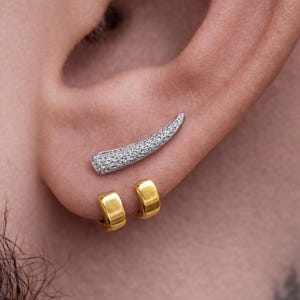Peut inclure: Gros plan de trois boucles d'oreilles sur une oreille. La boucle d'oreille supérieure est une barre incurvée argentée incrustée de petites pierres transparentes. En dessous, deux petites créoles dorées. Le teint est clair.
