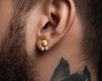 May include: A close-up of an ear with a gold-colored, circular earring. The earring is a flat disc with a brushed texture. Below the earring are two silver-colored, rectangular pieces. The person has a beard and a tattoo on their neck.