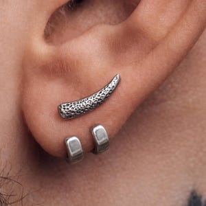 Puede incluir: Juego de joyas para la oreja en tono plateado. El conjunto incluye un trepador de oreja curvado y texturizado y dos pendientes de aro pequeños. El trepador de oreja tiene una superficie detallada y picada. Los pendientes se muestran en una oreja.
