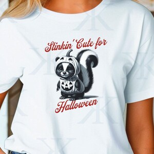 Può includere: Maglietta bianca con un'illustrazione in bianco e nero di una puzzola che indossa un costume da zucca. Il testo "Stinkin' Cute for Halloween" è stampato in rosso sulla maglietta.