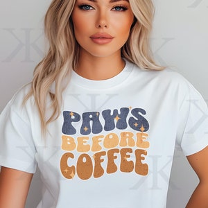 以下が含まれることがあります： ゴールドのグリッターのアクセントが付いた、ブルー、オレンジ、ブラウンのレトロなスタイルのグラフィックが書かれた白いTシャツ。グラフィックには「PAWS BEFORE COFFEE」と書いてあります。