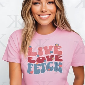 Może przedstawiać: Różowa koszulka z grafiką w stylu retro, na której widnieje napis "Live Love Fetch" w kolorowej czcionce z sercami.