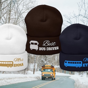 May include: Three beanies with bus graphics. One is white with "Mrs EMMA", another is brown with "Best BUS DRIVER", and the third is navy with "Mr KYLE". A yellow school bus drives on a snowy road in the background.