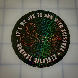 May include: A black and iridescent round sticker with the text "Athletic Trainer - It's My Job To Run With Scissors" in white lettering. The sticker features a pair of scissors in the center.