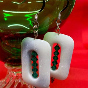 May include: White, rectangular earrings with a central cutout, filled with green beads, hang from silver hooks. The earrings are set against a red background, with a green glass bowl in the background.