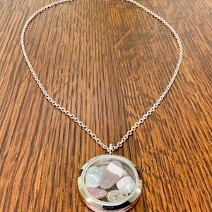 May include: Silver-toned necklace with a round, clear locket pendant. The pendant contains small, colorful stones. The necklace is displayed on a wooden surface.
