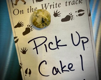 May include: A white note with black paw prints and the text "On the Write track" and "Pick Up Cake!" A green and white stone magnet is attached to the note.