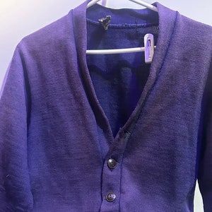 May include: A purple cardigan with a large embroidered patch on the left side. The patch features a stylised letter "C" and the number "59". The cardigan has a v-neck and five buttons down the front.