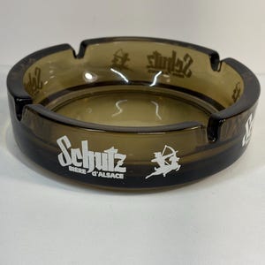 Pode incluir: Um cinzeiro castanho com um design circular. O cinzeiro tem uma borda larga com quatro entalhes para apoiar os cigarros. O texto "Schutz Biere d'Alsace" é impresso em branco, juntamente com um gráfico de um arqueiro.