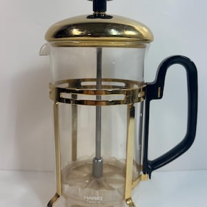 Puede incluir: Una cafetera de prensa francesa de vidrio transparente con detalles metálicos dorados y un asa negra. La tapa y la base tienen un acabado dorado. La marca "HARIO" y el modelo "THA1-4" están impresos en el cristal.
