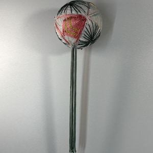 Puede incluir: Una bola decorativa japonesa hecha a mano de color blanco y verde con un diseño geométrico rosa y dorado. La bola está suspendida de una cuerda y tiene una larga borla verde.