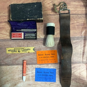 Peut inclure: Un kit de barbier vintage avec une courroie en cuir noir, un blaireau à raser, un porte-lames, une pierre à aiguiser en Carborundum, un crayon rouge et blanc rayé et deux cartes de prix bleues et orange qui indiquent "Rodney Barber Shop, J.B. Stinson, Prop. 12 Rasages pour 1 €."