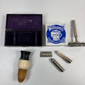 Peut inclure: Ensemble d'articles de rasage vintage, comprenant une boîte doublée de velours violet, une boîte de savon à raser Williams, un rasoir de sûreté, un blaireau et une lame de rasoir. Les articles sont sur une surface blanche.