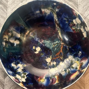 May include: A large, blue and white ceramic bowl with a floral design. The bowl has a deep, rounded shape and is decorated with a stylized tree branch with white flowers and leaves. The background is a deep blue with hints of purple and green.