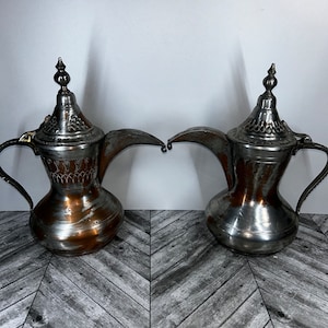Puede incluir: Dos cafeteras árabes tradicionales de plata y cobre con diseños ornamentados y asas. Las cafeteras están sobre una superficie gris con patrón de chevron.