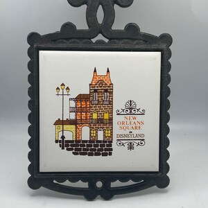 May include: A black cast iron trivet with a white ceramic tile featuring a colourful illustration of a building in New Orleans Square at Disneyland. The text "NEW ORLEANS SQUARE in DISNEYLAND" is printed on the tile.