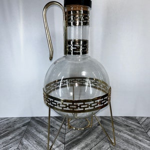 Può includere: Una caraffa di vetro trasparente con un supporto in metallo dorato e un tappo di sughero. La caraffa ha un corpo rotondo e un collo lungo con un manico. Il supporto ha tre gambe e una base rotonda.