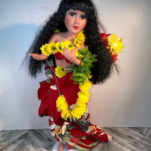 May include: A porcelain doll wearing a red and green dress with yellow flower leis. The doll has long black hair and brown eyes.
