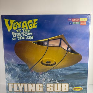 Pode incluir: Caixa de modelo do Flying Sub de "Viagem ao Fundo do Mar". A arte da caixa mostra um submarino amarelo emergindo da água azul. O texto inclui "Flying Sub" e "All Plastic Assembly Model Kit".