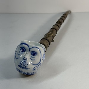 Puede incluir: Una pipa decorativa con un cuenco de cerámica blanca adornado con una cara de mono azul. El vástago es de metal con bandas decorativas. Un objeto de colección único, la pipa es una pieza decorativa.