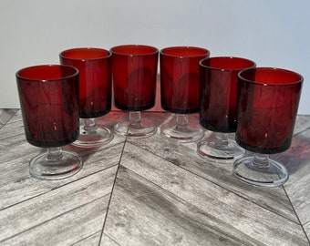May include: Set of six red glass shot glasses with clear glass stems and bases. The glasses are arranged in a semi-circle on a wood surface.