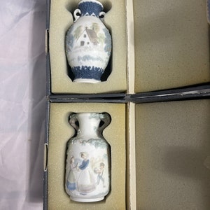 May include: Two small porcelain vases with intricate designs. The top vase features a white background with a blue and white house and trees. The bottom vase features a white background with a blue and white scene of people in a garden.