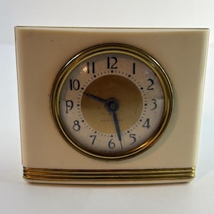 May include: A vintage cream-coloured rectangular clock with a gold-tone circular clock face. The clock face has black numerals and hands, with a gold-tone bezel. The clock has a gold-tone trim at the base and around the clock face.