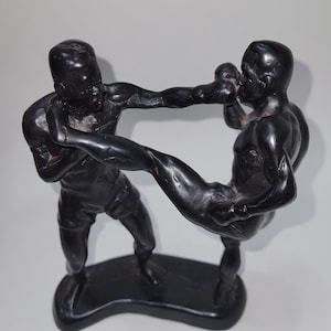 Könnte beinhalten: Schwarze und weiße Figur von zwei Figuren in Kampfpose. Die Figur links steht mit erhobenem rechten Bein, während die Figur rechts in Abwehrhaltung mit erhobenem linken Arm steht.