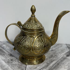 May include: A vintage brass teapot with detailed engraved patterns. The teapot has a curved spout, a rounded body, and a handle. The lid is topped with a decorative finial. The teapot is displayed on a wooden surface.