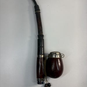 May include: A vintage tobacco pipe with a dark brown wooden stem and a curved black mouthpiece. A small, matching wooden container with a silver lid and a string with two wooden beads is next to the pipe.