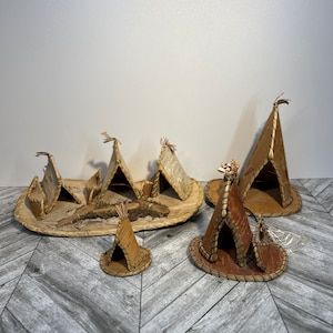 Puede incluir: Un conjunto de seis tipis en miniatura hechos de corteza de abedul y cuero. Los tipis están dispuestos en círculo sobre una base tejida. Los tipis están decorados con costuras y flecos. El conjunto incluye una etiqueta que dice "Artesanía india".