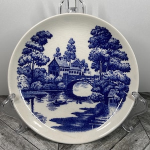 Puede incluir: Un plato de cerámica azul y blanco con una escena de cabaña. El plato presenta un puente sobre un río, árboles y un pequeño bote.