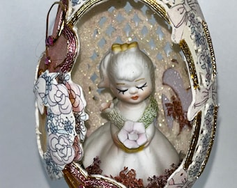 Puede incluir: Una delicada figurita de porcelana de una niña que sostiene una flor se exhibe dentro de una cáscara de huevo tallada y decorada. La cáscara de huevo está adornada con intrincados diseños florales y detalles dorados. La figurita está rodeada de flores rosas y blancas y purpurina.