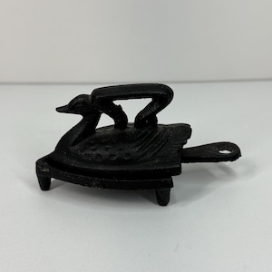 May include: A black cast iron trivet shaped like a swan with a textured body and a handle extending from the back. The swan figure is mounted on a flat base supported by three small legs, featuring a detailed wing pattern and a curved neck.