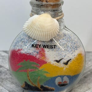 May include: A glass bottle filled with colourful sand depicting a palm tree, ocean, and birds. The bottle has a cork stopper and a seashell tied to the neck. The label on the bottle reads "KEY WEST".