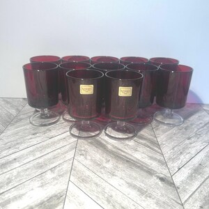 May include: Set of 12 clear glass stemmed shot glasses with a deep red hue. The glasses have a small base and a wide mouth. The glasses are labeled "Lumenart" in gold lettering.