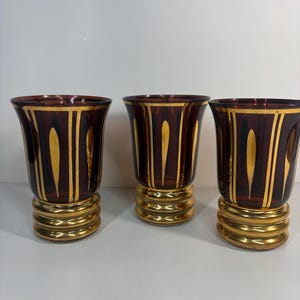 May include: Three burgundy-coloured drinking glasses with gold accents. Each glass has a gold rim, vertical gold stripes, and a gold base. The glasses are likely for decorative or formal use.