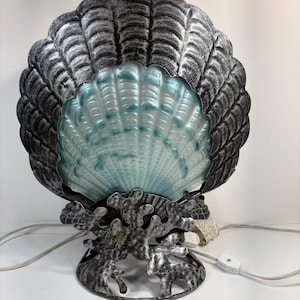 Peut inclure: Lampe décorative en forme de coquillage avec une finition argentée et noire. La lampe présente une coquille en verre bleu-vert et repose sur une base en forme de corail. La lampe a un cordon avec un interrupteur marche/arrêt.