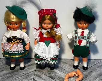 May include: Three traditional German dolls wearing colorful clothing. The dolls are standing on a wooden floor. One doll is wearing a green dress with a white apron and a red hat with white polka dots. Another doll is wearing a green dress with a white apron and a green hat with a white feather. The third doll is wearing a green shirt with a red tie and green pants.
