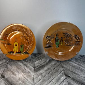 Könnte beinhalten: Zwei dekorative Keramikteller mit goldfarbenem Rand und braunem Hintergrund. Die Teller zeigen handgemalte Szenen von japanischen Frauen in traditioneller Kleidung mit Bäumen und Blumen.
