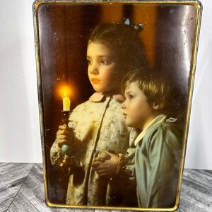 Puede incluir: Caja de hojalata vintage con una imagen de dos niños sosteniendo una vela. La niña lleva un vestido floral y el niño lleva una camisa azul. El fondo es de color marrón oscuro.