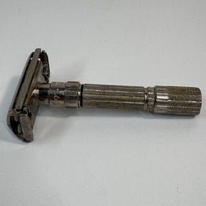 Puede incluir: Una maquinilla de afeitar de seguridad plateada vintage con un mango texturizado. El cabezal de la maquinilla es rectangular y el mango tiene un diseño estriado para un mejor agarre. La maquinilla está diseñada para afeitarse.