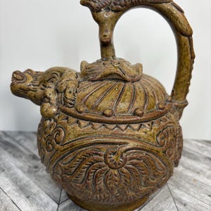 Puede incluir: Una tetera de cerámica marrón con intrincados tallados de hojas y flores. La tetera tiene un pico en forma de dragón y un asa en forma de pájaro.