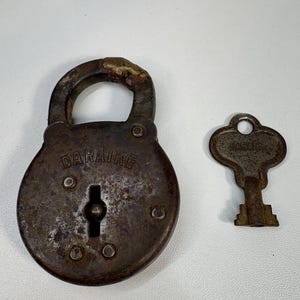 Puede incluir: Un candado y una llave oxidados y antiguos. El candado es de color marrón oscuro con la palabra "DARAIKE" grabada en la parte delantera. La llave es de color marrón oxidado a juego.