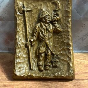 Puede incluir: Una placa de pared de madera con una imagen tallada de una persona que sostiene un bastón y una luna creciente. La placa tiene una superficie rugosa y texturizada y está pintada en tonos marrones.