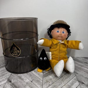 Peut inclure: Ensemble d'articles Patch Kids, comprenant un baril en plastique gris foncé avec un logo, une poupée en combinaison jaune et casquette marron, et une figurine en peluche noire avec des yeux blancs et des pieds jaunes.